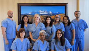 A group photo of ten dental clinic staff members in blue scrubs posing together in front of a scenic mountain landscape painting